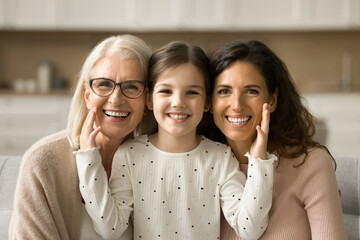 Cheerful cute little girl, mom, grandmother sitting close indoors, hugging, looking at camera with...