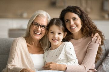 Happy mom and elderly grandma hugging cute tween girl, posing for family portrait on home couch,...