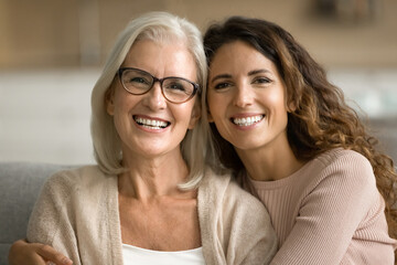 Happy blonde elderly mom and young daughter woman posing at home, looking at camera with toothy smiles, laughing, hugging, enjoying warm family relationship, bonding. Head shot portrait