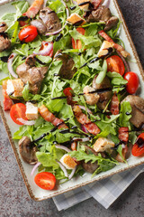 Warm salad with grilled chicken liver,  bacon, cherry tomatoes, arugula, red onion and croutons close-up on plate on table. Vertical top view from above