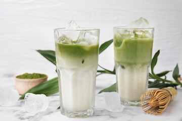 Glasses of tasty iced matcha latte, bamboo whisk and leaves on white marble table