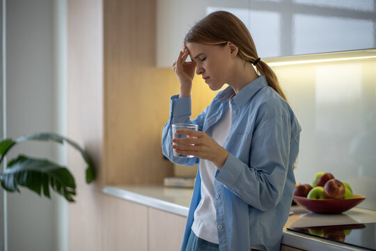 Woman feeling terrible pain in head drinking water on kitchen at home with closed eyes. Girl having headache, migraine, cephalalgia, premenstrual syndrome. Taking medications, painkillers concept.