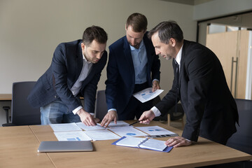 Three businessmen in formal suits negotiating, discuss joint project, working in teamwork, share ideas, review sales statistics, discuss marketing plan, take part in group meeting in conference room
