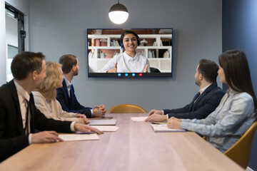 Indian woman leads on-line conference with diverse teammates sit at desk look at tv screen sit at desk in modern boardroom. HR manager interviewing Indian woman applicant answer questions at interview - obrazy, fototapety, plakaty