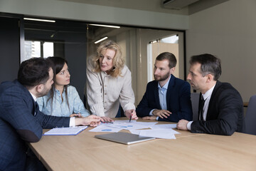 Group of millennial and middle-aged businesspeople take part in seminar led by mature female business coach share strategy, review together charts infographic, do paperwork gathered together in office