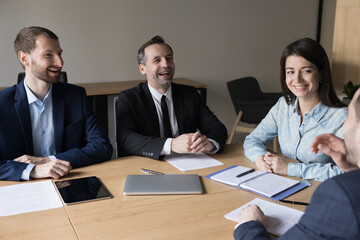 Group of smiling positive employees, businesspeople sit at desk solving current business at morning briefing, informal meeting, enjoy jokes, feel happy, friendly relationships and successful teamwork