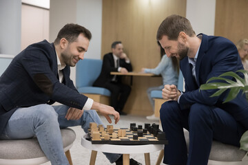 Two happy millennial 35s businessmen, office employees, colleagues have fun engaged in intellectual game, play chess during break in modern coworking space. Rest at workday, friendship, hobby activity