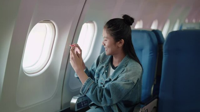 Asian female traveler capturing outside views through her smartphone's window on the airplane, eager for holiday adventures
