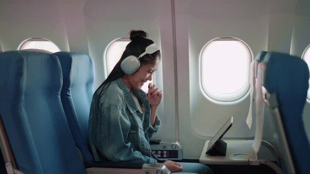 Asian female traveler enjoying a show on her tablet with headphones during the plane journey, anticipating holiday adventures.