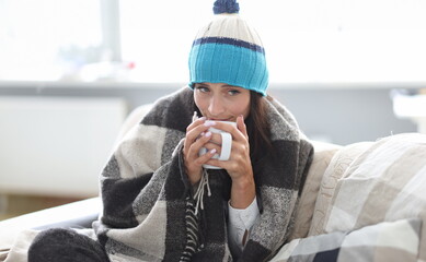 Beautiful woman in hat and plaid sit on sofa and hold white cup in hand. Sick person drink hot...