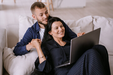 young married couple lying on couch with laptop looking memorable photos. young man with beard is lying with his wife holding her hand making video call talking to parents after moving to another city