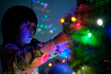 A 4-year-old charming little girl decorates a Christmas tree in light of garlands, her eyes sparkling with wonder as she hangs the ornaments.