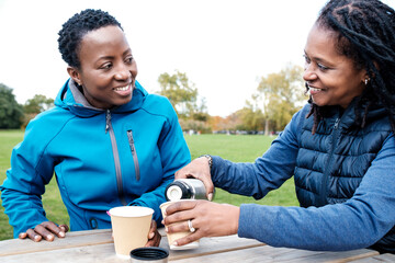 Two fitness friends pouring coffee on reusable cups outdoors.