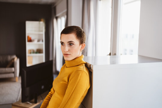 Young woman with short hair, wearing a yellow turtleneck, gazes sadly to the side in her living room.