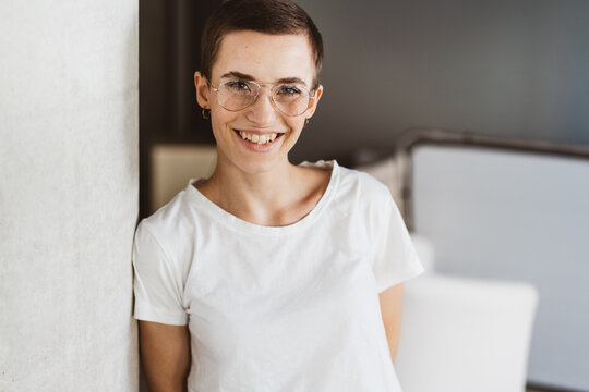 Joyful Young Woman with Short Hair, Glasses, and T-Shirt Laughing at Camera in Cozy Living Room