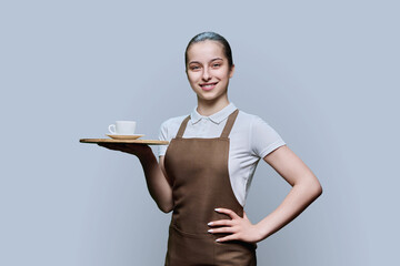 Young female waitress in apron with tray of cup of coffee on gray background
