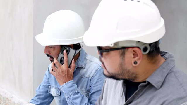 Closeup Asian two engineering male wearing hardhat using smartphone contact customer or inspector sitting in construction site, worker, architect, manager, business partner working with Indian labor