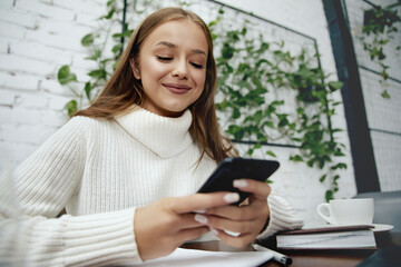 woman sitting at table studying using laptop reading a book. Girl takes a break holding mobile phone surfing internet.