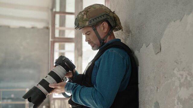 Caucasian male with grey beard leaning by wall and checking everything recorded on his professional camera. Working in dangerous location. Wearing safety precaution. Recording evidence.