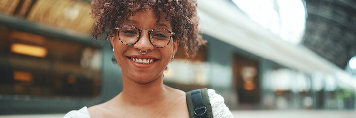 Close-up portrait of a young woman in glasses with a smartphone and backpack stands at the station...