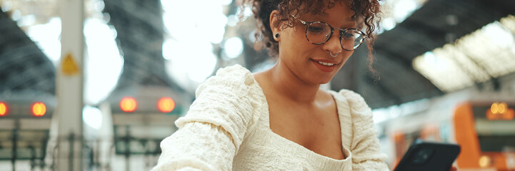 Close-up of a young woman in glasses sits at the station, looks into a smartphone and smiles....