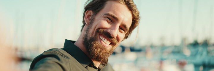 Closeup portrait of a smiling man with a beard chatting on the embankment, on a yacht background. Frontal close-up of happy young male hipster using mobile phone - obrazy, fototapety, plakaty