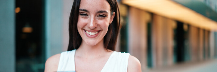 Beautiful woman with freckles and dark loose hair wearing white top is walking down the street with...