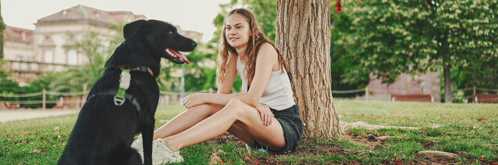 Cute girl with black dog is resting under tree on the lawn. Happy dog rejoices being on walk in the...