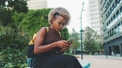 Smiling young woman in glasses, wearing brown top, uses mobile phone, chatting with friends on social networks while sitting outdoors