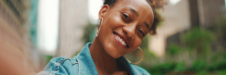 Closeup portret cute African girl with ponytail, wearing denim jacket makes stream, video call on smartphone in modern city background. - obrazy, fototapety, plakaty