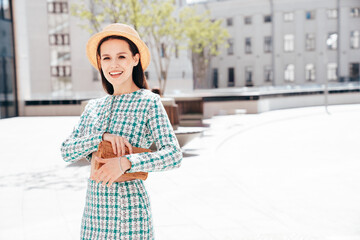 Young beautiful smiling woman in trendy summer green jacket and skirt clothes. Sexy carefree model posing in the street at sunset. Positive brunette female. Cheerful and happy. In hat, holds clutch