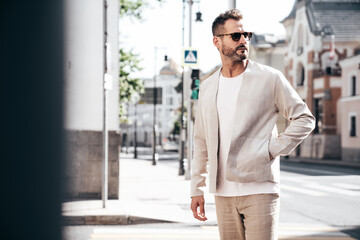 Handsome confident stylish hipster lambersexual model. Sexy modern man dressed in elegant white suit jacket. Fashion male posing in the street background in Europe city at sunset. In sunglasses