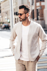 Obraz premium Handsome confident stylish hipster lambersexual model. Sexy modern man dressed in elegant white suit jacket. Fashion male posing in the street background in Europe city at sunset. In sunglasses