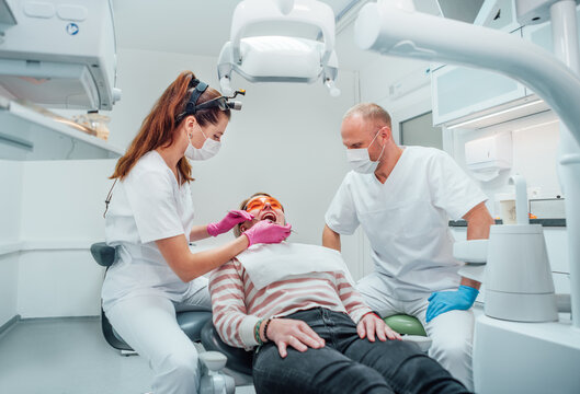 Dental clinic patient appointment. Dentist woman in magnifying glasses doing teeth prevention using medical tools. Young man assistant helping her. Health care and medicare industry concept image.