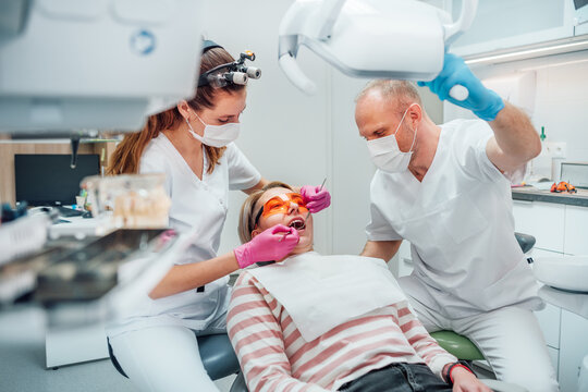 Dental clinic patient appointment. Dentist woman in magnifying glasses doing teeth prevention using medical tools. Young man assistant pointing light. Health care and medicare industry concept image.