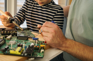 Father with his son spend time together. Close-up of a man teaching his boy at home to solder computer spare parts. Education moment during parenthood. Togetherness concept.