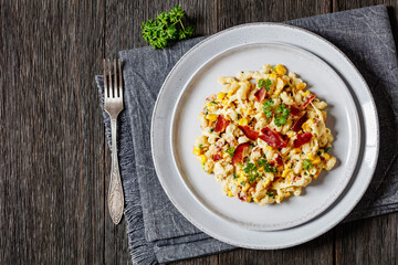 macaroni and cheese with corn, bacon topped with panko breadcrumbs on plate on dark wooden table with fork, horizontal view from above, flat lay, free space