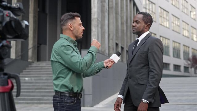 Handsome correspondent with microphone taking interview of handsome businessman or politician while standing in front of large office. Mixed-raced men actively communicating in front of camera.