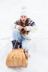 Fototapeta premium Selective focus vertical view of little blond girl in winter clothes making a funny face while sliding down a snowy slope in a vintage wooden sled