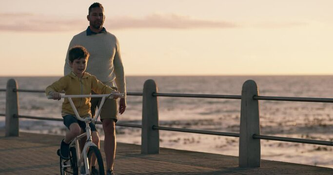 Father, child on bicycle and outdoor learning at beach with support, helping and bonding together on holiday. Parent, family and dad with boy or kid on bike for cycling and walking by sunset and sea