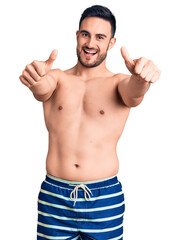 Fototapeta premium Young handsome man wearing swimwear approving doing positive gesture with hand, thumbs up smiling and happy for success. winner gesture.