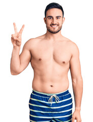 Fototapeta premium Young handsome man wearing swimwear smiling looking to the camera showing fingers doing victory sign. number two.