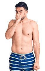 Fototapeta premium Young handsome man wearing swimwear smelling something stinky and disgusting, intolerable smell, holding breath with fingers on nose. bad smell