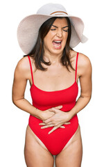 Beautiful brunette woman wearing swimsuit and summer hat with hand on stomach because nausea,...