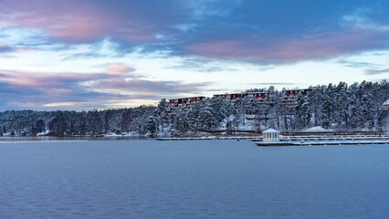 Winter snowy landscape of Gustavsberg, Sweden. Sunset clouds. The shore of Baltic sea bay overgrown with pines and firs covered with snow. Houses surrounded by forest. Scandinavia in winter season.