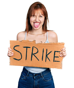 Redhead young woman holding strike banner cardboard sticking tongue out happy with funny expression.