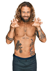 Handsome man with beard and long hair standing shirtless showing tattoos smiling funny doing claw...