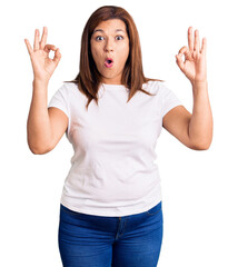 Middle age latin woman wearing casual white tshirt looking surprised and shocked doing ok approval symbol with fingers. crazy expression