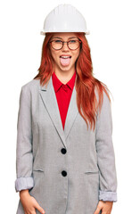 Young redhead woman wearing architect hardhat sticking tongue out happy with funny expression. emotion concept.