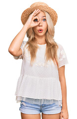 Young blonde girl wearing summer hat doing ok gesture shocked with surprised face, eye looking through fingers. unbelieving expression.